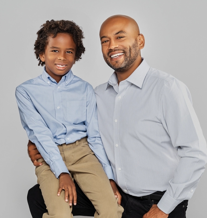 Matthew Gillian, candidate for Fresno City Council in 2023, is pictured here with his son. Matthew is on the right in the picture. He is a handsome Black man with a bald haircut and a trimmed beard. He is wearing a light blue shirt and black pants. Matthew is smiling. His son is sitting on his father's knee on the left side of the image. Matthew's son looks like he is about 7 years old. He is also Black and has short curly hair. He is wearing a light blue shirt and khaki pants. End of photo description.